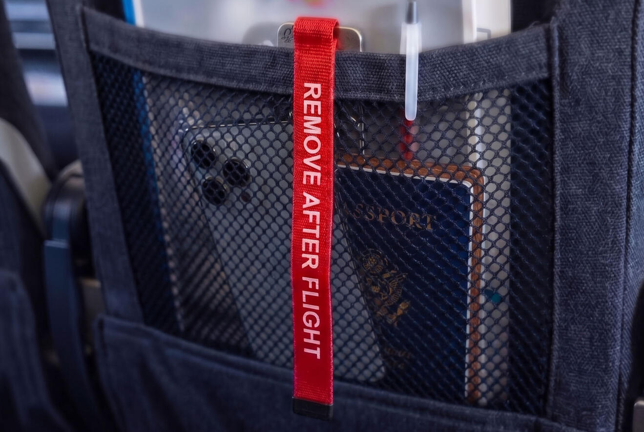 Red 'Remove After Flight' Streamer in Seat Pocket A close-up view from a passenger's perspective looking down into the fabric seatback pocket of an aircraft seat. The red 'REMOVE AFTER FLIGHT' streamer tag dangles visibly from the top edge of the pocket. Travel documents and a pen are partially visible in